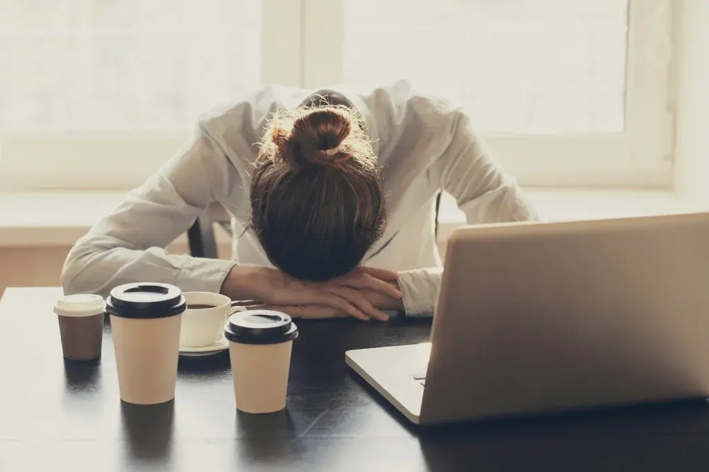 A person resting their head on folded arms at a desk beside a laptop and several coffee cups